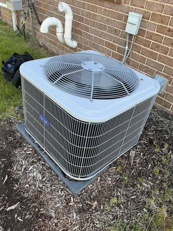 Completed Gold AC Inspection on a 6-year-old air conditioning system with condenser unit located on exterior concrete pad against brick wall and evaporator coil in basement. Performed comprehensive maintenance including visual inspection of outdoor condenser unit, verified proper positioning on concrete pad with adequate clearance, inspected cabinet housing and protective grilles for structural integrity, and confirmed fan guard assembly secure. Checked condensate drainage system including white PVC piping configuration mounted to exterior wall, verified electrical disconnect box properly installed and accessible. Inspected condenser coil fins through louvers, assessed unit for signs of wear, corrosion, or damage. Evaluated surrounding area including mulch bedding and confirmed no debris obstruction to airflow. Unit operating within normal parameters with clean metal surfaces and no visible deficiencies noted. System demonstrates proper installation standards and maintenance condition 