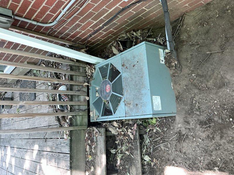 Responded to service call for insufficient airflow from 10+ year old air conditioning system with outdoor condenser unit located in basement area. Upon arrival, performed visual inspection of exterior condenser unit which revealed significant accumulation of leaves, organic debris, and dirt on and around equipment. Unit casing showed extensive weathering and age-related deterioration. Removed top panel to access internal components and assess operational status.

Discovered heavy debris buildup beneath and around condenser unit, including dead leaves and decomposed plant matter that had accumulated between unit and building foundation. Identified potential airflow restriction issues due to debris accumulation on fan grille and surrounding areas. Wooden mounting platform showed advanced deterioration from prolonged weather exposure.

Documented equipment condition through photographs showing exterior unit placement, internal component access, debris accumulation, and overall installatio