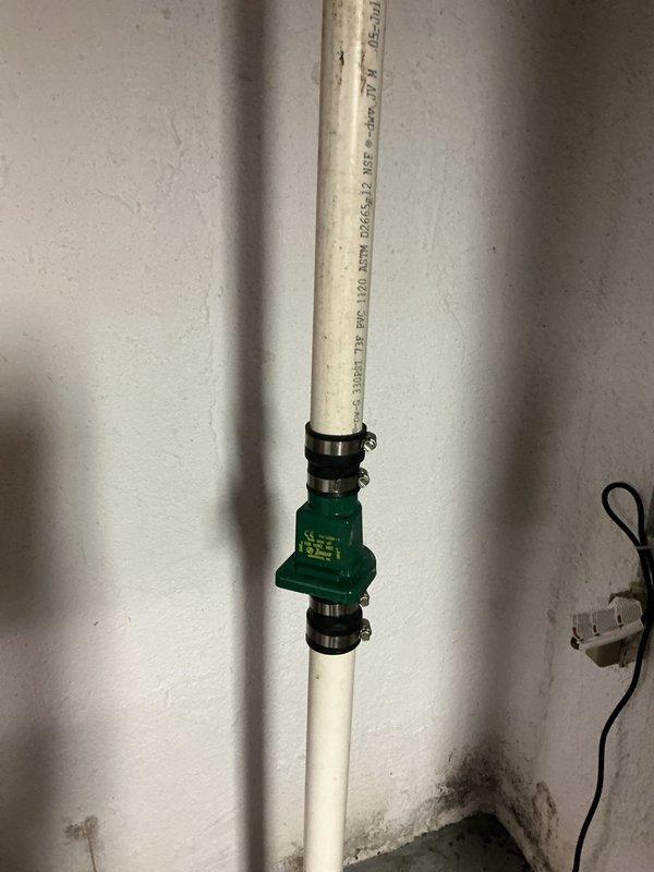 Responded to service call for reported sump pump leak. Upon inspection, identified leak at check valve assembly on discharge line. The green inline check valve showed signs of deterioration at connection points with visible moisture present. Evaluated entire discharge system including interior PVC piping and exterior drainage line. Verified proper operation of check valve mechanism and assessed seal integrity at upper and lower connection points. Inspected exterior discharge point where corrugated drainage pipe extends along building foundation, confirming proper water flow path away from structure. Documented system configuration including check valve installation, pipe routing, and exterior drainage setup. Photographed interior check valve assembly and exterior discharge termination point for service records. Provided homeowner with assessment findings and recommendations for addressing the leak source.