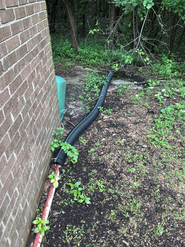 Responded to service call for reported sump pump leak. Upon inspection, identified leak at check valve assembly on discharge line. The green inline check valve showed signs of deterioration at connection points with visible moisture present. Evaluated entire discharge system including interior PVC piping and exterior drainage line. Verified proper operation of check valve mechanism and assessed seal integrity at upper and lower connection points. Inspected exterior discharge point where corrugated drainage pipe extends along building foundation, confirming proper water flow path away from structure. Documented system configuration including check valve installation, pipe routing, and exterior drainage setup. Photographed interior check valve assembly and exterior discharge termination point for service records. Provided homeowner with assessment findings and recommendations for addressing the leak source.