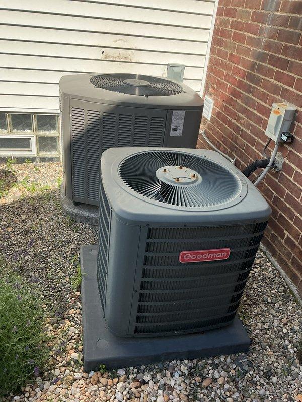 Completed comprehensive Gold AC inspection on dual-unit residential cooling system, both units approximately 3 years old with air handler/coil located in basement. Performed thorough evaluation of both outdoor condenser units, including Goodman brand unit, verifying proper operation, clearances, and ventilation access. Both systems confirmed operational with condensers properly positioned on concrete pads with adequate gravel drainage, clean exterior casings, unobstructed fan assemblies, and appropriate clearance from building structure for optimal airflow and serviceability.