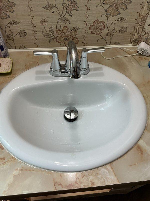 Completed installation of chrome two-handle centerset faucet assembly on bathroom sink. Installed new faucet with curved spout, conical handles, and pop-up drain stopper mechanism, connecting supply lines to existing copper plumbing beneath oval drop-in basin. Verified proper water flow, tested drain operation, and confirmed all connections were secure with no leaks detected.