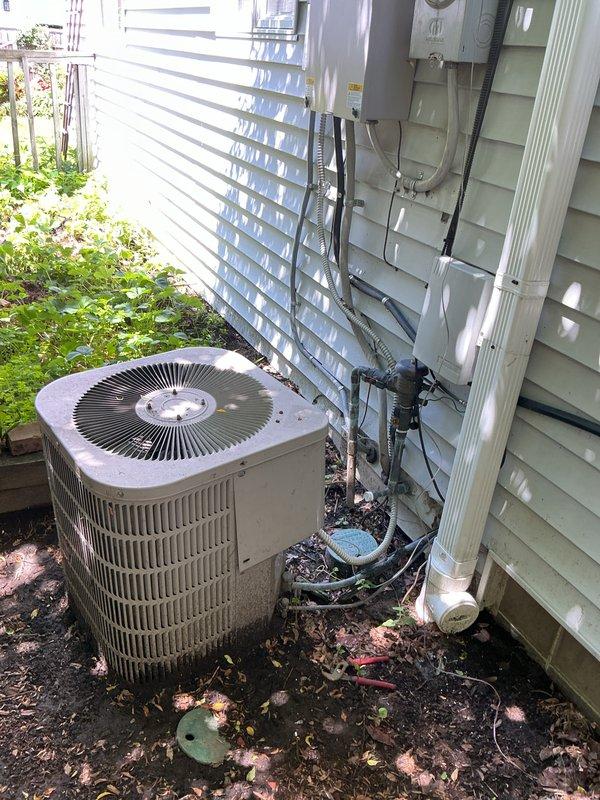 Conducted on-site evaluation for HVAC system replacement estimate including natural gas furnace, air conditioning unit, and mini-split system. Existing outdoor condensing unit located at ground level on building exterior with light green vinyl siding showed signs of weathering but remains operational. Documented current equipment configuration including electrical conduit routing, service panel connections, and refrigerant line placement. Assessed installation conditions noting accumulated debris around outdoor unit and existing mounting location on dark mulch base. Evaluated basement furnace location and upper floor equipment placement for replacement specifications. Photographed existing condensing unit showing cylindrical metal casing with vertical grille fins, top-mounted fan assembly with radial blade configuration, and surrounding electrical infrastructure. Reviewed natural gas fuel supply for furnace replacement requirements and confirmed accessibility for future installation wo