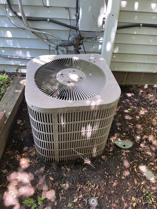 Conducted on-site evaluation for HVAC system replacement estimate including natural gas furnace, air conditioning unit, and mini-split system. Existing outdoor condensing unit located at ground level on building exterior with light green vinyl siding showed signs of weathering but remains operational. Documented current equipment configuration including electrical conduit routing, service panel connections, and refrigerant line placement. Assessed installation conditions noting accumulated debris around outdoor unit and existing mounting location on dark mulch base. Evaluated basement furnace location and upper floor equipment placement for replacement specifications. Photographed existing condensing unit showing cylindrical metal casing with vertical grille fins, top-mounted fan assembly with radial blade configuration, and surrounding electrical infrastructure. Reviewed natural gas fuel supply for furnace replacement requirements and confirmed accessibility for future installation wo