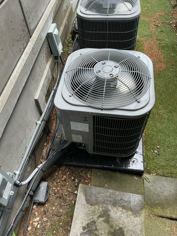 Completed Gold AC Inspection on two 8-year-old outdoor condensing units serving a multi-zone residential system with air handler located in basement. Performed comprehensive maintenance including visual inspection of both condenser units positioned on concrete pad, verified proper operation of fan motors and compressors, inspected condenser coils and fins for damage or blockage, checked electrical connections and disconnect boxes, confirmed refrigerant line integrity, and assessed overall system performance. Both units operating within normal parameters with adequate airflow through top-mounted fan grilles. Noted minor debris accumulation around unit perimeter and between condensers; area surrounding equipment shows typical seasonal leaf fall but no significant obstructions affecting operation. Electrical conduit and junction boxes properly secured to exterior wall with no visible damage. Units mounted on appropriate pads with proper clearances maintained. System confirmed operational 