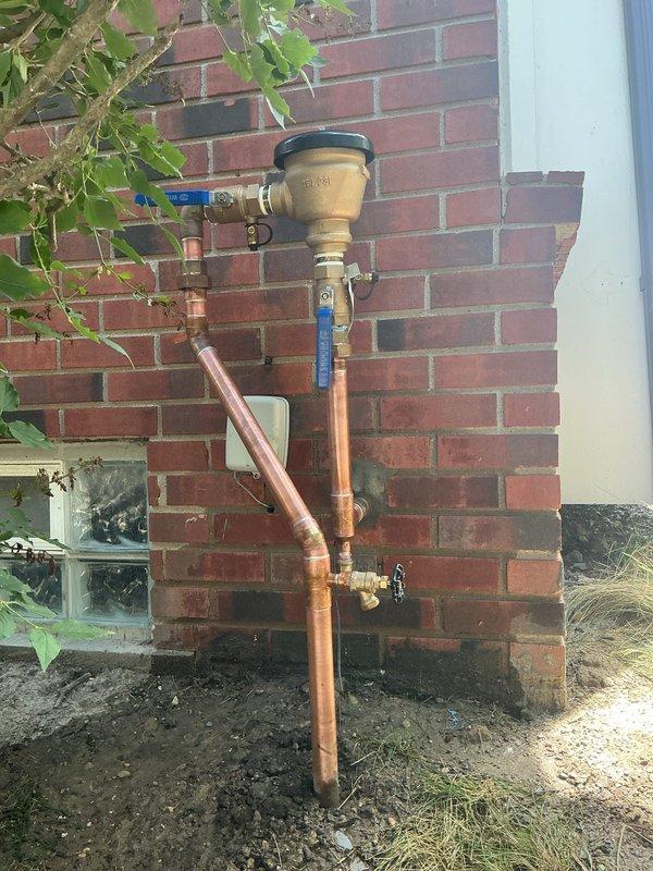 Installed a new 1 1/4" backflow preventer assembly on exterior brick wall using copper piping configuration with shutoff valves. Completed installation per permit requirements, ensuring proper vertical mounting and secure connections to existing plumbing system. Documented completed installation showing backflow device with test cocks, blue-handled shutoff valve at top, and black-handled isolation valve at bottom connection point.