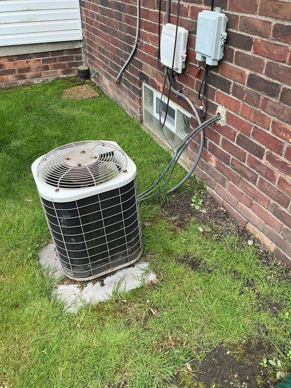 Responded to no cooling complaint affecting entire residence. Upon arrival, found aging cylindrical R-22 air conditioning condenser unit located at rear of property with unknown service history. System evaluation confirmed unit was operating with low refrigerant charge and utilizing obsolete R-22 refrigerant. Due to age of equipment, phase-out of R-22 refrigerant, and overall condition of condenser, provided comprehensive replacement estimate for new cooling system. Documented existing installation including outdoor unit placement, electrical service routing with conduit and disconnect boxes mounted to brick exterior, and current system configuration for replacement planning purposes.