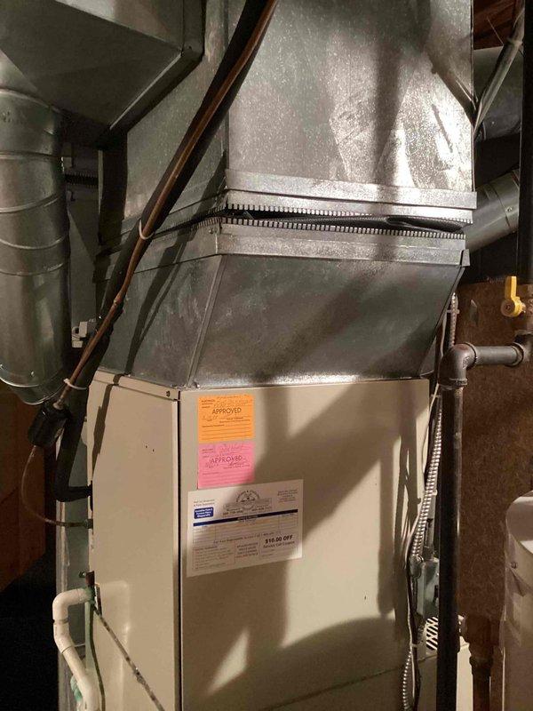 Completed estimate for replacement of 27-year-old furnace unit located in basement utility room. Assessed existing beige furnace installation with connected galvanized plenum and ductwork system, noting copper refrigerant lines and flexible metal conduit. Documented significant condensation buildup on existing ductwork throughout mechanical space, indicating potential efficiency concerns with aging system. Photographed current HVAC configuration including duct connections, structural mounting, and proximity to water heater and utility systems to provide comprehensive replacement specifications.
