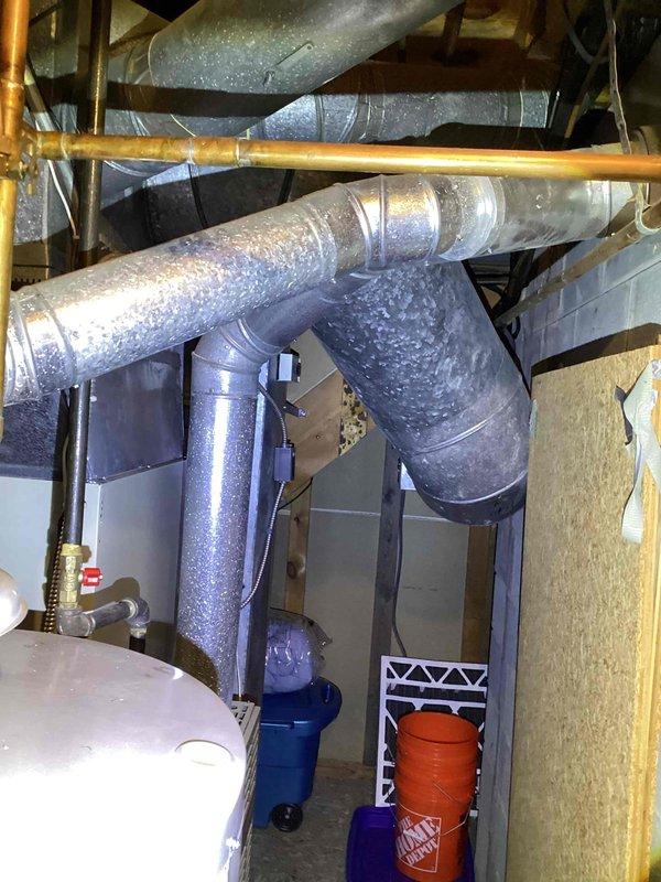 Completed estimate for replacement of 27-year-old furnace unit located in basement utility room. Assessed existing beige furnace installation with connected galvanized plenum and ductwork system, noting copper refrigerant lines and flexible metal conduit. Documented significant condensation buildup on existing ductwork throughout mechanical space, indicating potential efficiency concerns with aging system. Photographed current HVAC configuration including duct connections, structural mounting, and proximity to water heater and utility systems to provide comprehensive replacement specifications.