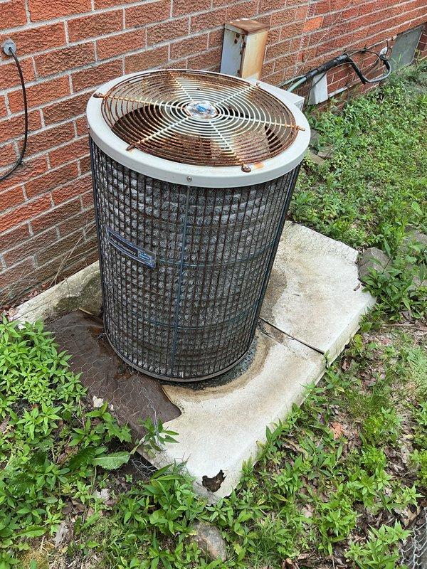 Completed on-site assessment of existing outdoor air conditioning condenser unit for replacement estimate. Unit exhibited significant weathering, rust, and oxidation throughout metal components and housing, with yellowed and deteriorated top cover indicating extended service life. Inspected electrical disconnect box, refrigerant line set with insulation, and conduit runs along brick exterior wall. Documented equipment condition, installation configuration, and utility connections. Photographed existing system components including concrete pad foundation with vegetation overgrowth. Provided comprehensive replacement estimate for new AC system installation.