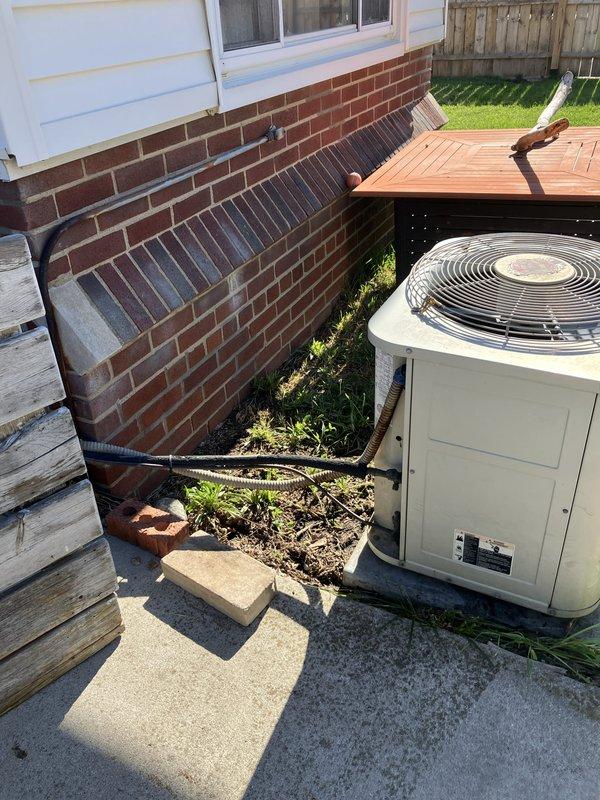 Completed estimate for replacement of 20-year-old residential air conditioning system located in backyard on concrete pad adjacent to house foundation. Evaluated existing beige condenser unit with top-mounted fan grille and assessed refrigerant line routing along brick foundation to basement furnace location. Documented current equipment condition and provided replacement options for aging AC system that has reached end of expected service life.