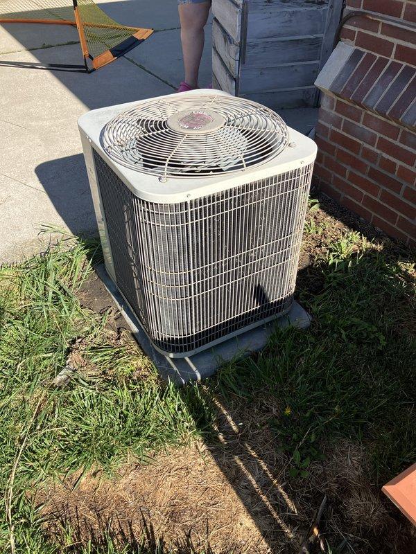 Completed estimate for replacement of 20-year-old residential air conditioning system located in backyard on concrete pad adjacent to house foundation. Evaluated existing beige condenser unit with top-mounted fan grille and assessed refrigerant line routing along brick foundation to basement furnace location. Documented current equipment condition and provided replacement options for aging AC system that has reached end of expected service life.