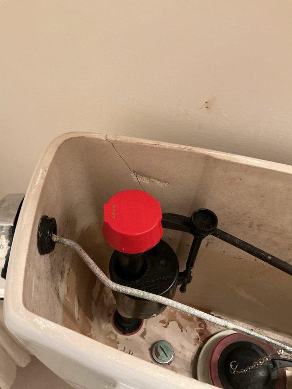 Inspected residential toilet system and documented existing conditions. Tank interior revealed significant mineral deposits, hard water staining, and a visible crack in the upper left portion of the ceramic tank wall. Fill valve assembly with float mechanism and flush valve with rubber flapper were operational but showing age-related wear consistent with the deteriorated tank condition. Photographed both exterior toilet installation and internal tank components for service documentation and recommended replacement due to structural compromise of the tank.