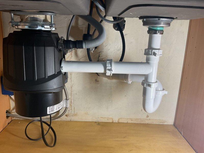 Completed installation of garbage disposal unit under double-basin kitchen sink with proper electrical connection and drain configuration. Installed white PVC P-trap assembly with secure threaded connections to accommodate disposal discharge and secondary sink drain. Verified proper operation of disposal unit, tested all drain connections for leaks, and ensured proper venting of the drainage system.