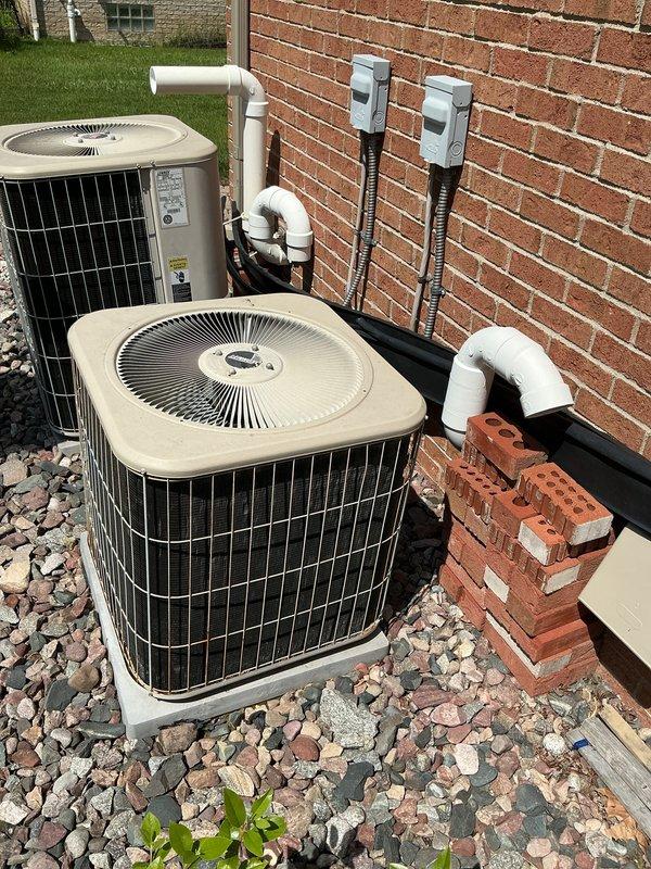 Completed full replacement of 20+ year-old outdoor air conditioning system with new condenser unit installed on gravel pad against exterior brick wall. New installation includes proper electrical disconnect boxes mounted to wall with conduit runs, PVC refrigerant line connections, and dual-unit configuration for enhanced cooling capacity. System tested and operational upon completion.