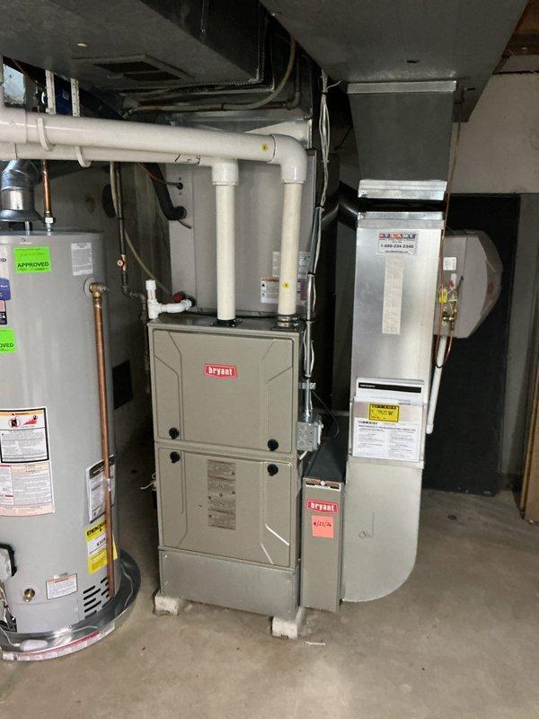 Completed comprehensive Gold AC Inspection on Bryant residential air conditioning system. Performed full system evaluation including outdoor condenser unit positioned on concrete pad against building exterior and indoor air handler cabinet located in mechanical room alongside Bryant furnace and water heater. Conducted standard tune-up procedures including visual inspection of condenser coil, fan operation verification, electrical connection assessment, and refrigerant line examination. Verified proper clearances around outdoor unit, checked mounting stability on concrete pad, and inspected electrical conduit connections from building to unit. Evaluated indoor components including ductwork connections and air handler operation. System operational at time of inspection with no immediate repairs required. Unit age unknown per service request. Documented equipment condition through photographic records showing installation configuration and component placement for future service reference.