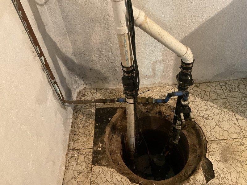 Provided estimate for sump pump upgrade in basement of 53-year-old home. Assessed existing sump pump system with failed water-powered backup that experienced mineral deposit buildup. Evaluated current installation including primary pump, discharge piping with PVC and copper components, check valves, and electrical connections. Discussed battery backup system options to replace water backup due to previous failure and concerns about power outages from recent storms. Photographed existing setup showing vertical discharge piping with shut-off valve, electrical conduit, sump pit with active water level, and tile floor installation around pit opening. Presented upgrade recommendations for battery-operated backup sump pump system to provide reliable secondary pumping capacity during power failures without dependency on municipal water pressure.