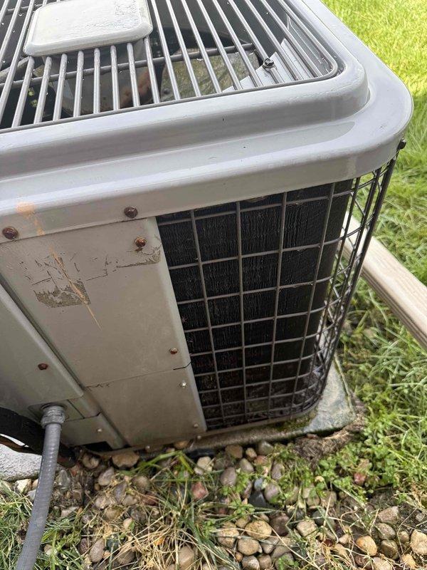 Performed comprehensive inspection and tune-up on 27-year-old residential air conditioning system located in backyard. Evaluated condenser unit which exhibited significant rust corrosion on exterior cabinet panels and fastener points, indicating advanced age and weathering. Measured supply air temperature at 96.9°F using calibrated digital thermometer at ductwork. Inspected condenser coils through front grille mesh, assessed refrigerant lines and electrical conduit connections. Examined thermostat operation and verified proper communication with cooling system. Conducted visual inspection of humidifier components and hot water tank as part of complete home comfort system evaluation. Unit showed signs of considerable deterioration consistent with its age, including rust staining, paint degradation, and general wear on cabinet surfaces. Provided assessment of remaining equipment lifespan based on observed condition of condenser unit, internal components, and overall system performance. A