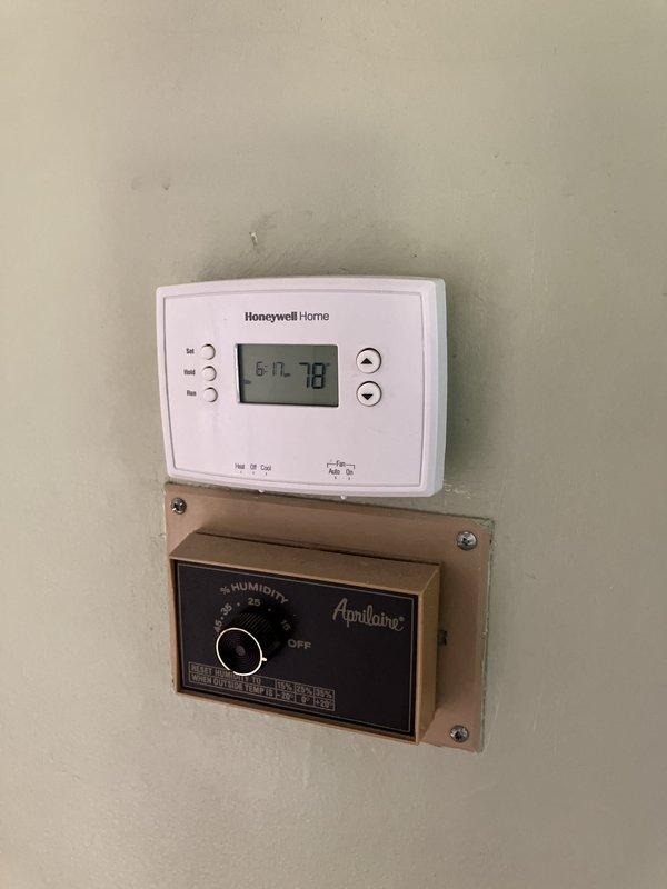 Completed estimate for replacement of aging outdoor air conditioning condenser unit currently utilizing obsolete R22 refrigerant. The existing beige metal-grilled unit showed visible weathering and age-related deterioration, positioned on grass against vinyl siding with standard electrical conduit installation. Assessment included evaluation of indoor climate control system featuring Honeywell Home digital thermostat and Aprilaire humidistat for compatibility with replacement equipment recommendations.