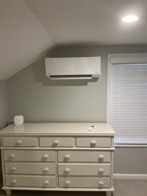 Completed full mini-split system installation including indoor wall-mounted evaporator unit in upper-level bedroom and outdoor condenser unit mounted on exterior brick wall in side yard. Ran refrigerant line set and electrical connections between indoor and outdoor components. Installed new dedicated electrical circuit to power the system. Indoor unit positioned on wall above dresser for optimal air distribution in sloped ceiling space. Outdoor condenser secured to brick wall with mounting brackets on gravel pad in side yard alongside existing HVAC equipment. System tested and verified operational upon completion.