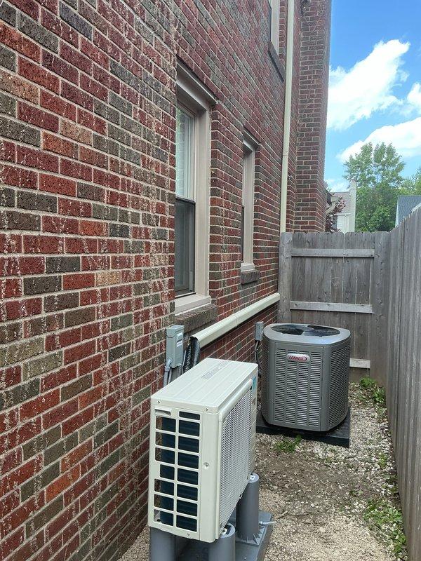 Completed full mini-split system installation including indoor wall-mounted evaporator unit in upper-level bedroom and outdoor condenser unit mounted on exterior brick wall in side yard. Ran refrigerant line set and electrical connections between indoor and outdoor components. Installed new dedicated electrical circuit to power the system. Indoor unit positioned on wall above dresser for optimal air distribution in sloped ceiling space. Outdoor condenser secured to brick wall with mounting brackets on gravel pad in side yard alongside existing HVAC equipment. System tested and verified operational upon completion.
