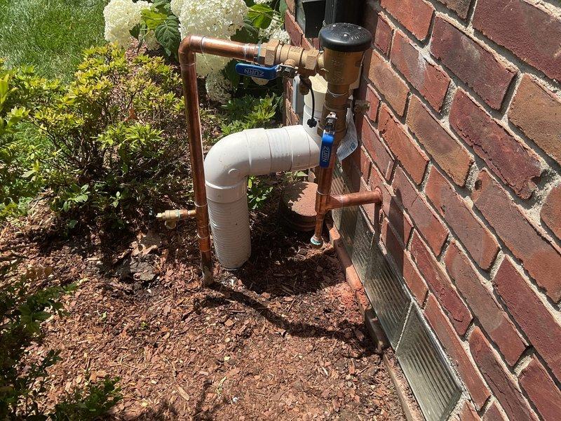 Completed installation of PVB (Pressure Vacuum Breaker) backflow prevention device on exterior residential water supply line. Integrated new backflow preventer assembly with existing copper piping system, utilizing proper PVC connections and shut-off valves for code compliance. Installation mounted securely to brick exterior wall with appropriate clearances for testing and maintenance access, positioned above grade level in landscaped area to prevent submersion and ensure proper drainage.