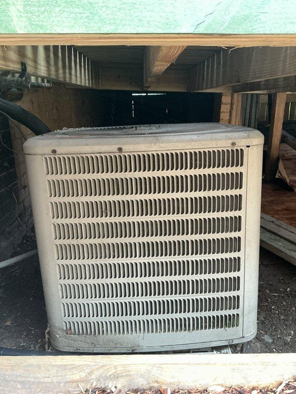 Responded to no cooling complaint on 2012 air conditioning system with outdoor condenser unit located beneath deck structure. Upon inspection, found unit blowing warm air and performed diagnostic evaluation of electrical connections, refrigerant levels, and compressor operation. Documented equipment condition and installation location in basement area with photos showing condenser placement and associated electrical components including junction box and conduit routing.