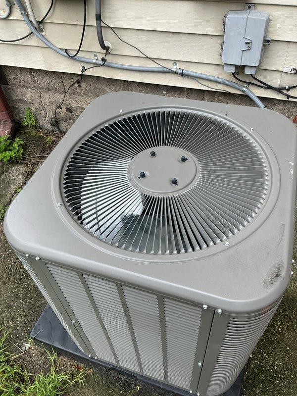 Performed diagnostic inspection on 3-year-old residential air conditioning system located in basement. Inspected outdoor condenser unit positioned on concrete pad adjacent to home exterior, confirming proper airflow through vertical louvers and radial fan grille assembly. Verified electrical connections at junction box and conduit routing along exterior wall. Examined indoor air handler ventilation grille and confirmed proper louver operation. Unit found in operational condition with parts under manufacturer warranty coverage.