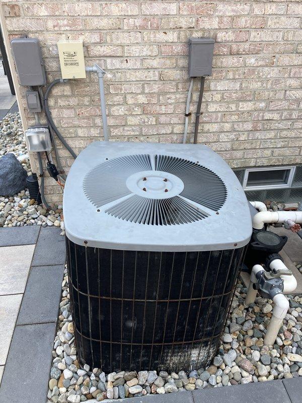 Conducted comprehensive site evaluation for high-efficiency HVAC system replacement. Assessed existing outdoor condenser unit with circular radial ventilation pattern, documented current electrical infrastructure including multiple meter housings and junction boxes mounted on exterior brick wall, and photographed refrigerant line routing and electrical disconnect components. Evaluated equipment placement, existing river rock landscaping base, and electrical service configuration to develop full system replacement estimate specifications.