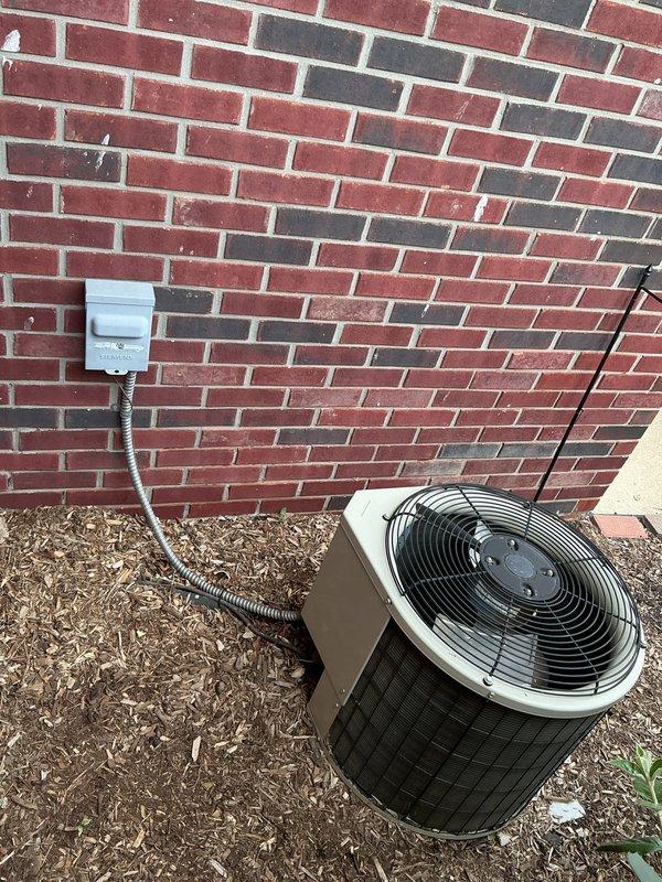Conducted on-site evaluation of existing outdoor air conditioning condenser unit located in exterior side yard between brick walls for equipment replacement estimate. Unit is a cylindrical design with top-mounted fan assembly, electrical disconnect box with conduit connection, and positioned on mulch bed against residential brick structure. Documented current equipment configuration, electrical service routing, installation clearances, and access constraints to provide comprehensive replacement system specifications and installation requirements.