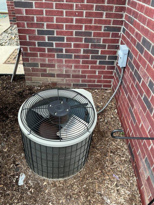 Conducted on-site evaluation of existing outdoor air conditioning condenser unit located in exterior side yard between brick walls for equipment replacement estimate. Unit is a cylindrical design with top-mounted fan assembly, electrical disconnect box with conduit connection, and positioned on mulch bed against residential brick structure. Documented current equipment configuration, electrical service routing, installation clearances, and access constraints to provide comprehensive replacement system specifications and installation requirements.