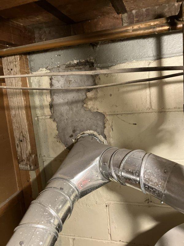 Conducted furnace replacement estimate for 25-year-old heating system located in basement. Inspected existing furnace installation, photographed current equipment configuration including white metal furnace unit with top-mounted access panel, PVC venting, flexible aluminum ductwork, and associated copper piping. Documented basement conditions showing concrete block walls, exposed floor joists, and utility space layout. Assessed ductwork connections including flexible ribbed ducting entering through concrete wall opening and rigid metal sections. Evaluated existing gas lines, condensate drains, and ventilation system components. No current operational issues reported by homeowner at time of inspection. Provided equipment replacement recommendations based on age of existing system and current installation configuration.