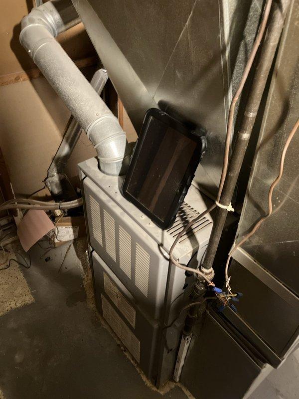 Conducted furnace replacement estimate for 25-year-old heating system located in basement. Inspected existing furnace installation, photographed current equipment configuration including white metal furnace unit with top-mounted access panel, PVC venting, flexible aluminum ductwork, and associated copper piping. Documented basement conditions showing concrete block walls, exposed floor joists, and utility space layout. Assessed ductwork connections including flexible ribbed ducting entering through concrete wall opening and rigid metal sections. Evaluated existing gas lines, condensate drains, and ventilation system components. No current operational issues reported by homeowner at time of inspection. Provided equipment replacement recommendations based on age of existing system and current installation configuration.