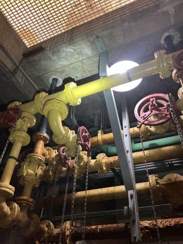 Performed commercial backflow prevention device testing on single unit as requested. Upon arrival, accessed mechanical room and located backflow assembly within overhead piping system featuring yellow and green painted pipes with multiple valve configurations and brass fittings. Conducted standard testing procedures on the backflow preventer, verified proper operation of check valves and relief valve, and documented system condition with photographic evidence showing the installation environment and equipment location within the facility's plumbing infrastructure.