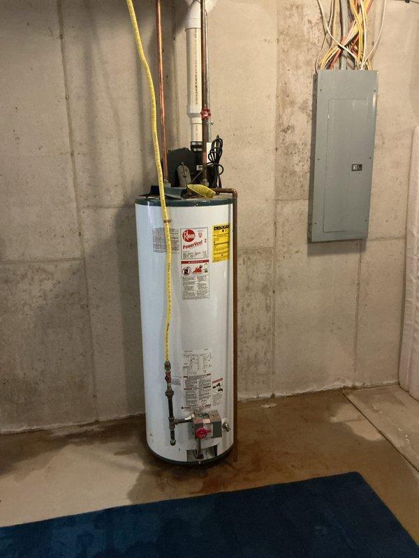 Responded to service call for leaking water heater in basement utility area. Upon arrival, performed visual inspection of existing white cylindrical tanked water heater unit. Confirmed active leak with visible water staining and discoloration present on concrete floor surrounding base of unit. Documented current installation including copper supply piping, yellow flexible gas line connection, and electrical connections. Assessed overall condition of water heater, noting manufacturer labels and safety warnings intact. Photographed unit from multiple angles to document leak location, plumbing configuration, and surrounding mechanical room conditions including proximity to electrical panel. Provided homeowner with professional assessment and replacement options for failed water heater unit.