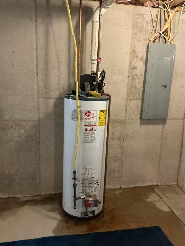 Responded to service call for leaking water heater in basement utility area. Upon arrival, performed visual inspection of existing white cylindrical tanked water heater unit. Confirmed active leak with visible water staining and discoloration present on concrete floor surrounding base of unit. Documented current installation including copper supply piping, yellow flexible gas line connection, and electrical connections. Assessed overall condition of water heater, noting manufacturer labels and safety warnings intact. Photographed unit from multiple angles to document leak location, plumbing configuration, and surrounding mechanical room conditions including proximity to electrical panel. Provided homeowner with professional assessment and replacement options for failed water heater unit.