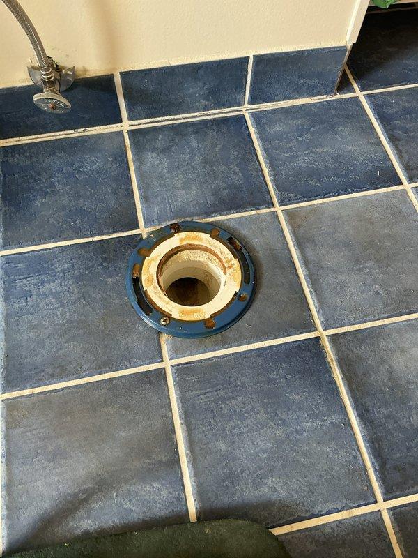 Completed plumbing estimate for toilet replacement on second-floor bathroom. Assessed existing white two-piece toilet installation on blue tile flooring and documented conditions including floor drain fixture with visible mineral deposits and rust staining. Noted heavy toilet unit requiring two-technician installation due to weight and second-floor location, with existing unit currently positioned on first floor awaiting transport and installation.