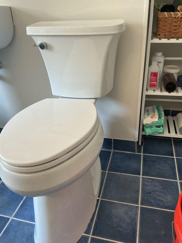 Completed plumbing estimate for toilet replacement on second-floor bathroom. Assessed existing white two-piece toilet installation on blue tile flooring and documented conditions including floor drain fixture with visible mineral deposits and rust staining. Noted heavy toilet unit requiring two-technician installation due to weight and second-floor location, with existing unit currently positioned on first floor awaiting transport and installation.