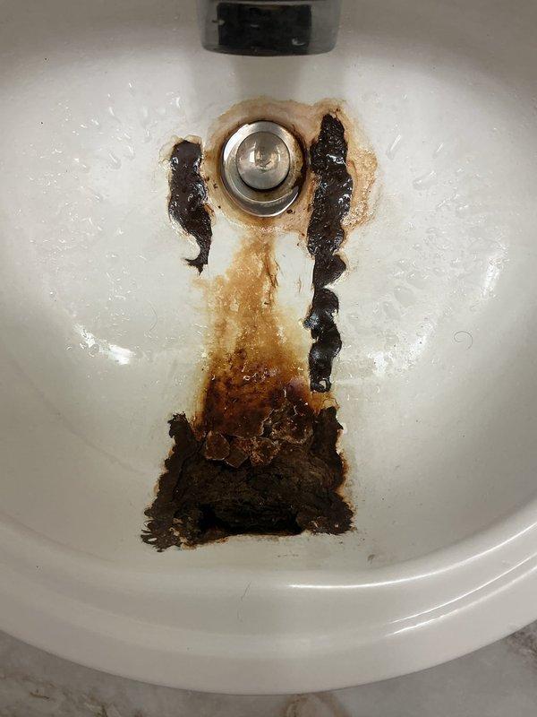 Completed on-site assessment of main floor bathroom plumbing requiring fixture and drain line replacement. Documented extensive deterioration of existing porcelain sink showing severe rust staining, mineral deposits, and discoloration concentrated around drain assembly. Inspected undersink plumbing revealing heavily corroded metal drain pipes with advanced rust damage throughout vertical and horizontal runs. Noted deteriorated pipe insulation and compromised connection points indicating long-term moisture exposure and aging infrastructure. Photographed all affected components including sink basin condition and corroded drain assembly to provide comprehensive documentation for replacement estimate. Recommended complete removal and replacement of sink fixture and associated drain piping due to extent of corrosion and potential for imminent failure.