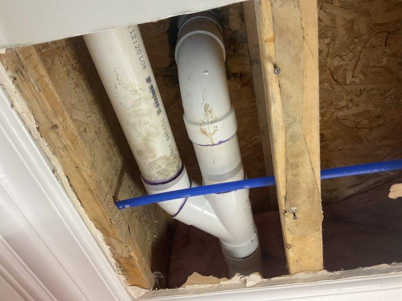 Responded to water leak originating from ceiling area caused by toilet flange failure. Accessed exposed ceiling cavity to inspect PVC drainage piping and toilet waste connection. Found compromised flange assembly with evidence of water infiltration around toilet drain connection. Evaluated existing 3-inch PVC waste line configuration, including elbows and straight sections with visible purple primer markings. Documented piping system condition showing dirt accumulation and staining on pipe exteriors. Identified drainage system routing through floor joists with intersecting PEX supply lines. Recommended flange replacement and proper sealing to prevent future water intrusion into ceiling space below.