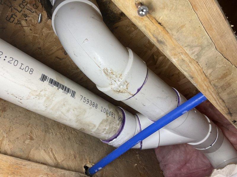 Responded to water leak originating from ceiling area caused by toilet flange failure. Accessed exposed ceiling cavity to inspect PVC drainage piping and toilet waste connection. Found compromised flange assembly with evidence of water infiltration around toilet drain connection. Evaluated existing 3-inch PVC waste line configuration, including elbows and straight sections with visible purple primer markings. Documented piping system condition showing dirt accumulation and staining on pipe exteriors. Identified drainage system routing through floor joists with intersecting PEX supply lines. Recommended flange replacement and proper sealing to prevent future water intrusion into ceiling space below.