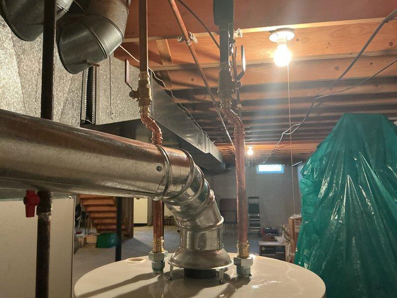 Completed installation of a 50-gallon Bradford White tanked water heater in basement utility area. Unit was properly positioned on concrete floor with all required plumbing connections, gas line hookup, and metallic vent pipe system installed to code, extending through ceiling joists with appropriate elbow fittings for proper exhaust ventilation.
