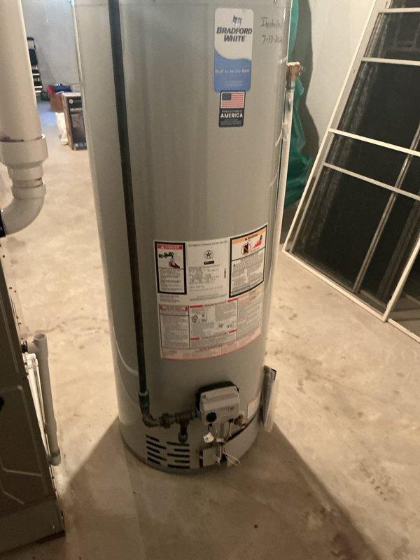 Completed installation of a 50-gallon Bradford White tanked water heater in basement utility area. Unit was properly positioned on concrete floor with all required plumbing connections, gas line hookup, and metallic vent pipe system installed to code, extending through ceiling joists with appropriate elbow fittings for proper exhaust ventilation.
