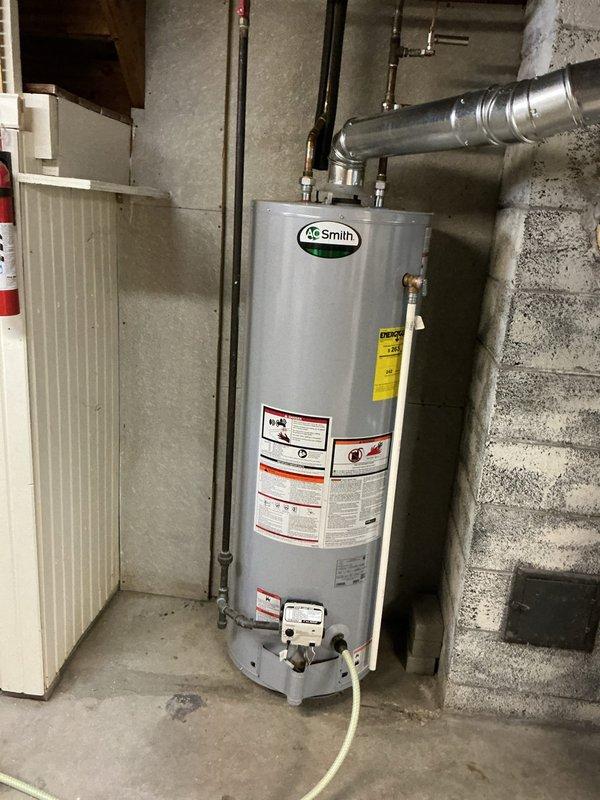 Responded to no hot water complaint in a 10-year-old residential property. Upon inspection, found an AO Smith tanked water heater in the basement with complete loss of hot water supply throughout the home. Documented existing installation with copper supply piping, brass shut-off valves, and proper venting configuration. Assessed unit condition, verified all safety labels and warning stickers were present and legible, and confirmed water heater was properly positioned on concrete floor against concrete block wall with appropriate clearances. Photographed the installation showing piping connections at top of unit including red and yellow-handled valves, metallic ductwork, and overall utility room setup for service records.