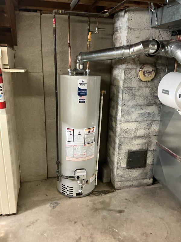 Responded to no hot water complaint in a 10-year-old residential property. Upon inspection, found an AO Smith tanked water heater in the basement with complete loss of hot water supply throughout the home. Documented existing installation with copper supply piping, brass shut-off valves, and proper venting configuration. Assessed unit condition, verified all safety labels and warning stickers were present and legible, and confirmed water heater was properly positioned on concrete floor against concrete block wall with appropriate clearances. Photographed the installation showing piping connections at top of unit including red and yellow-handled valves, metallic ductwork, and overall utility room setup for service records.