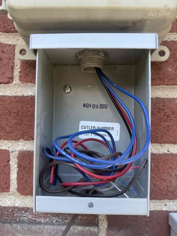 Responded to no cooling call on 13-year-old Bryant air conditioning system. Upon arrival, confirmed blower motor operating but condenser unit not running and no cooling to structure. Inspected outdoor unit and electrical connections at exterior junction box mounted on brick wall near condenser. Documented existing wiring configuration and Cutler-Hammer electrical components with installation date of November 2012. Photographed Comverge load control device and associated wiring connections for diagnostic purposes. System requires further electrical troubleshooting to determine root cause of condenser failure and restore cooling operation.