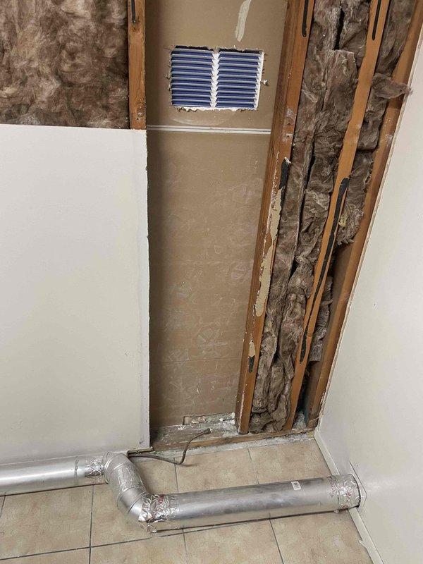 Provided estimate for replacement of 40-year-old natural gas wall furnace unit located in garage/laundry room. Existing unit is operational but has reached end of service life. Documented current installation including exposed wall cavity with deteriorated backing panel, flexible ductwork configuration on tile floor, and gas line connections with threaded fittings showing oxidation and age-related wear.