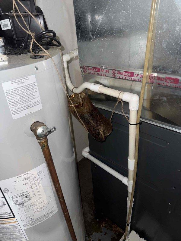 Responded to service call for water leak near furnace with 10-year-old AC system still providing cooling. Upon inspection, identified water pooling in basement mechanical room originating from condensate drainage system. Examined PVC condensate drain lines, fittings, and connections throughout the HVAC equipment area, noting accumulated debris and standard wear on piping components. Photographed condensate drain configuration including white PVC piping with elbow fittings, brass valve assembly, and connection points near ductwork and water heater to document existing conditions and drainage pathway.