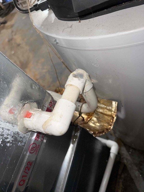Responded to service call for water leak near furnace with 10-year-old AC system still providing cooling. Upon inspection, identified water pooling in basement mechanical room originating from condensate drainage system. Examined PVC condensate drain lines, fittings, and connections throughout the HVAC equipment area, noting accumulated debris and standard wear on piping components. Photographed condensate drain configuration including white PVC piping with elbow fittings, brass valve assembly, and connection points near ductwork and water heater to document existing conditions and drainage pathway.
