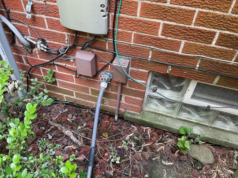 Responded to severe leak at exterior faucet mounted on back wall beneath kitchen window where water infiltration had begun entering the home and causing dirt accumulation on interior windowsill. Inspected outdoor spigot installation on 60-year-old brick structure near generator and assessed water meter assembly in basement access area showing significant staining on concrete base, indicating chronic moisture issues. Documented exterior wall conditions including utility connections, PVC drainage, glass block basement window, and foundation plantings, as well as interior meter installation with copper supply lines and shut-off valves to determine source and extent of leak for repair recommendations.